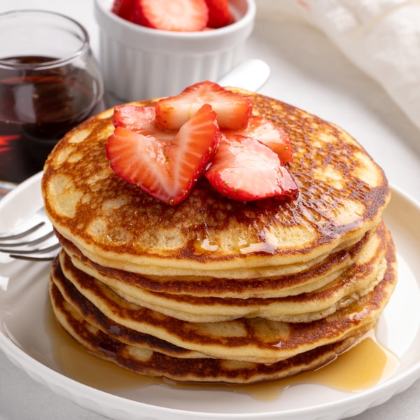 Closeup of image of sliced strawberries top a stack of golden-brown pancakes with syrup running over the pancakes and onto a white plate.
