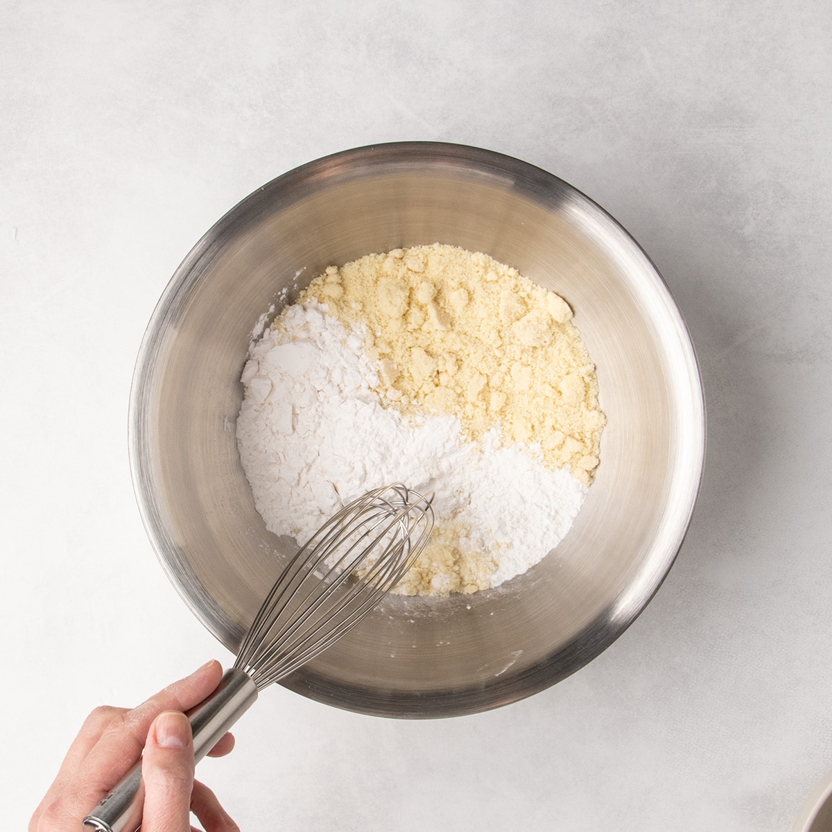 A hand holds a whisk over separate piles of white and pale brown dry ingredients in a stainless steel mixing bowl.