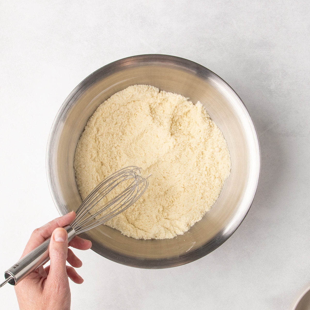 A hand holds a whisk over pale brown dry ingredients combined in a stainless steel mixing bowl.