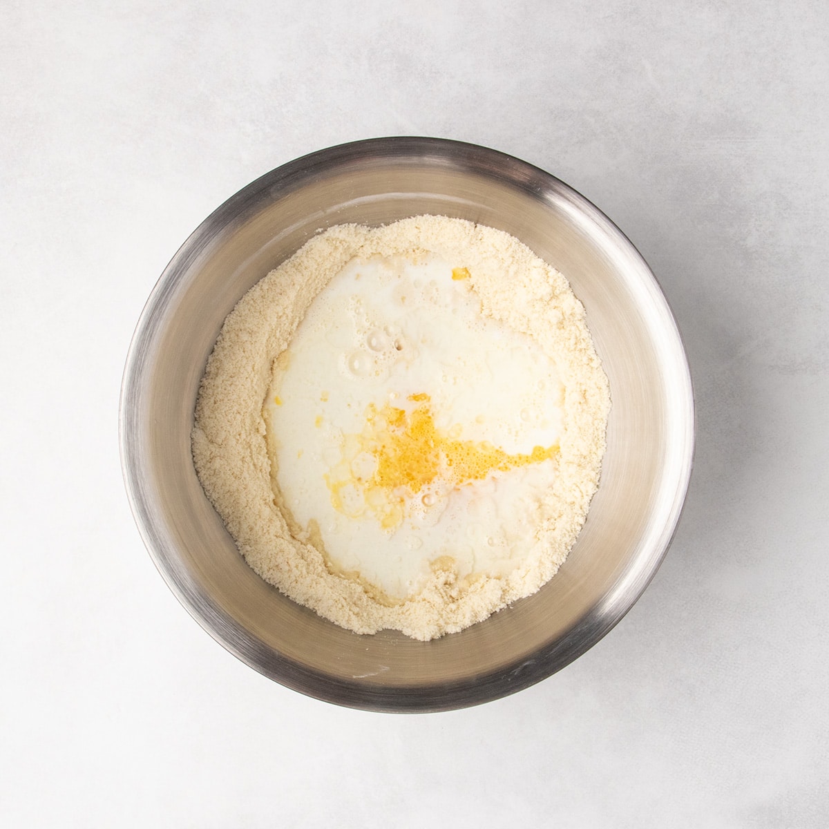White liquid and yellow liquid sit in the center of the pale brown dry ingredients in a stainless steel mixing bowl.