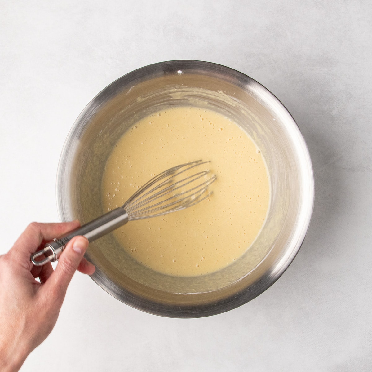 A hand holds a whisk in a stainless steel bowl filled with pale light brown batter, smooth except for some small bubbles from mixing.