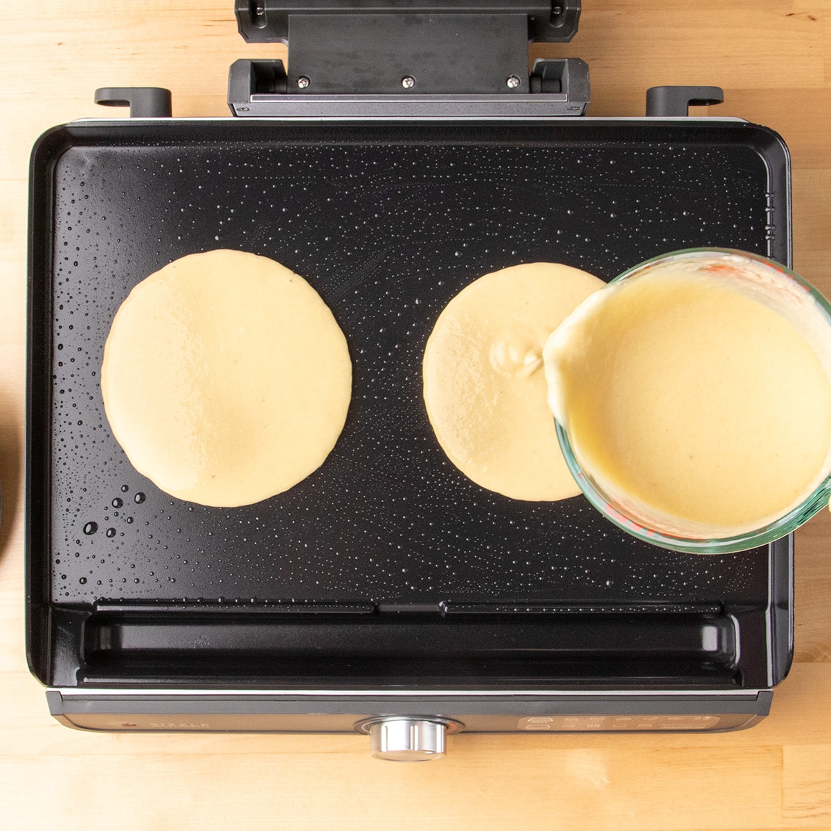 Two rounds of pale batter cook on an oiled black griddle as more batter is poured from a glass measuring cup.