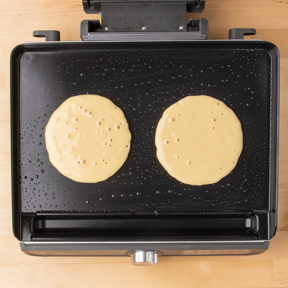 Large bubbles form on the surfaces of two pancakes cooking on an oiled black griddle.