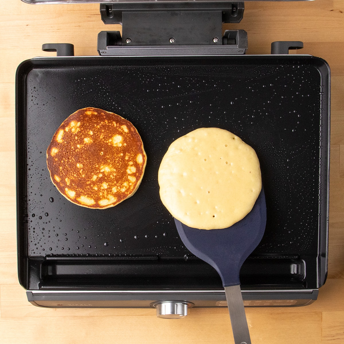 A spatula lifts one pale brown pancake beside a golden browned pancake on a black griddle.