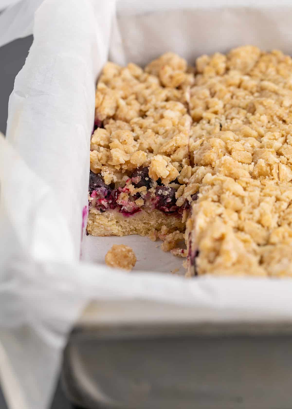 Pan of blueberry bars topped with crumble with one bar missing in metal pan with white liner