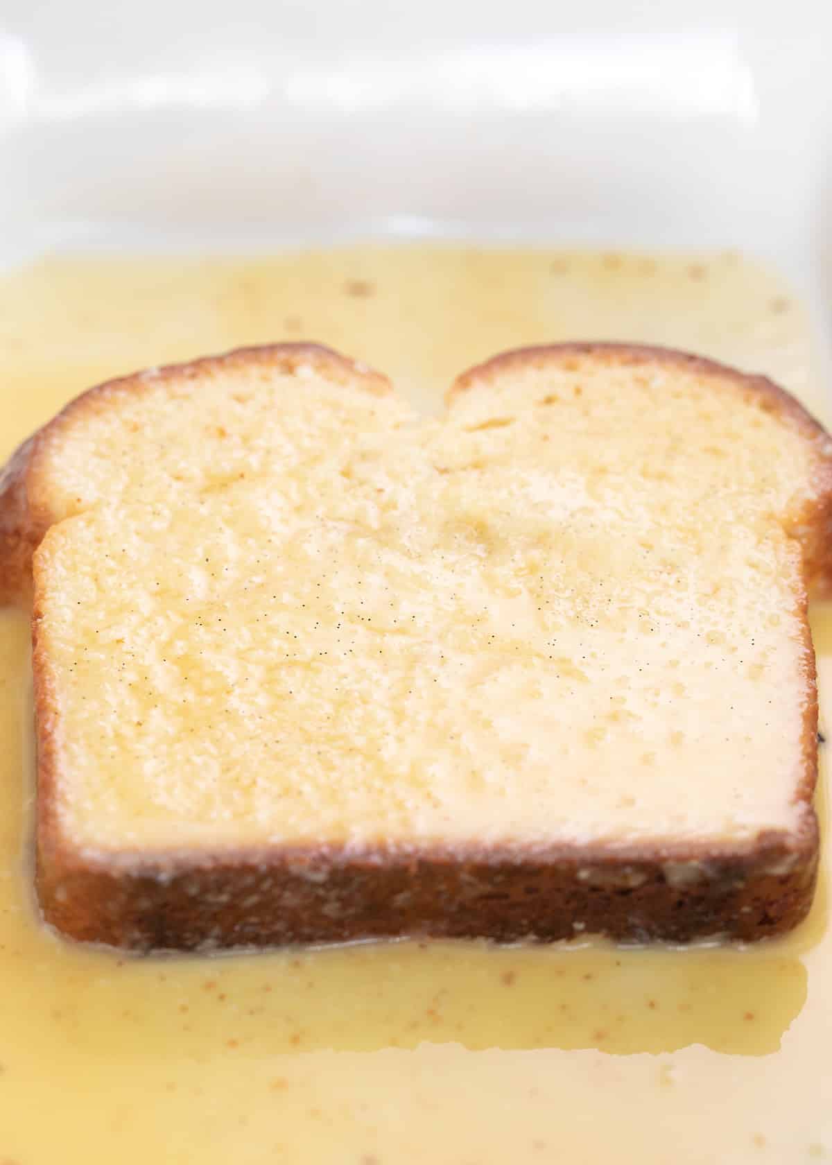 Slice of brioche bread soaking in egg mixture in white casserole dish