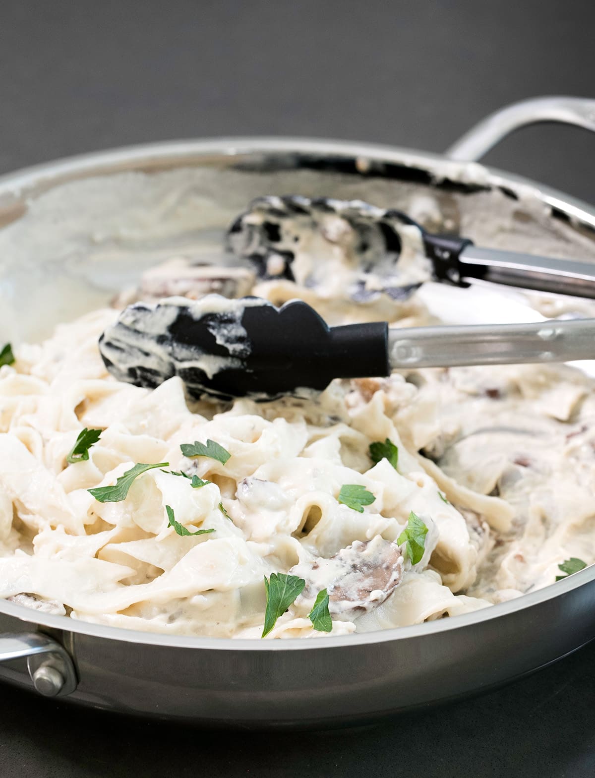 A pair of tongs serves the sauce and pasta from a metal skillet.