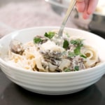 closeup of fingers twirling a forkful of vegan alfredo sauce on fettuccine pasta with sliced mushrooms and green parsley in a wide flat white bowl