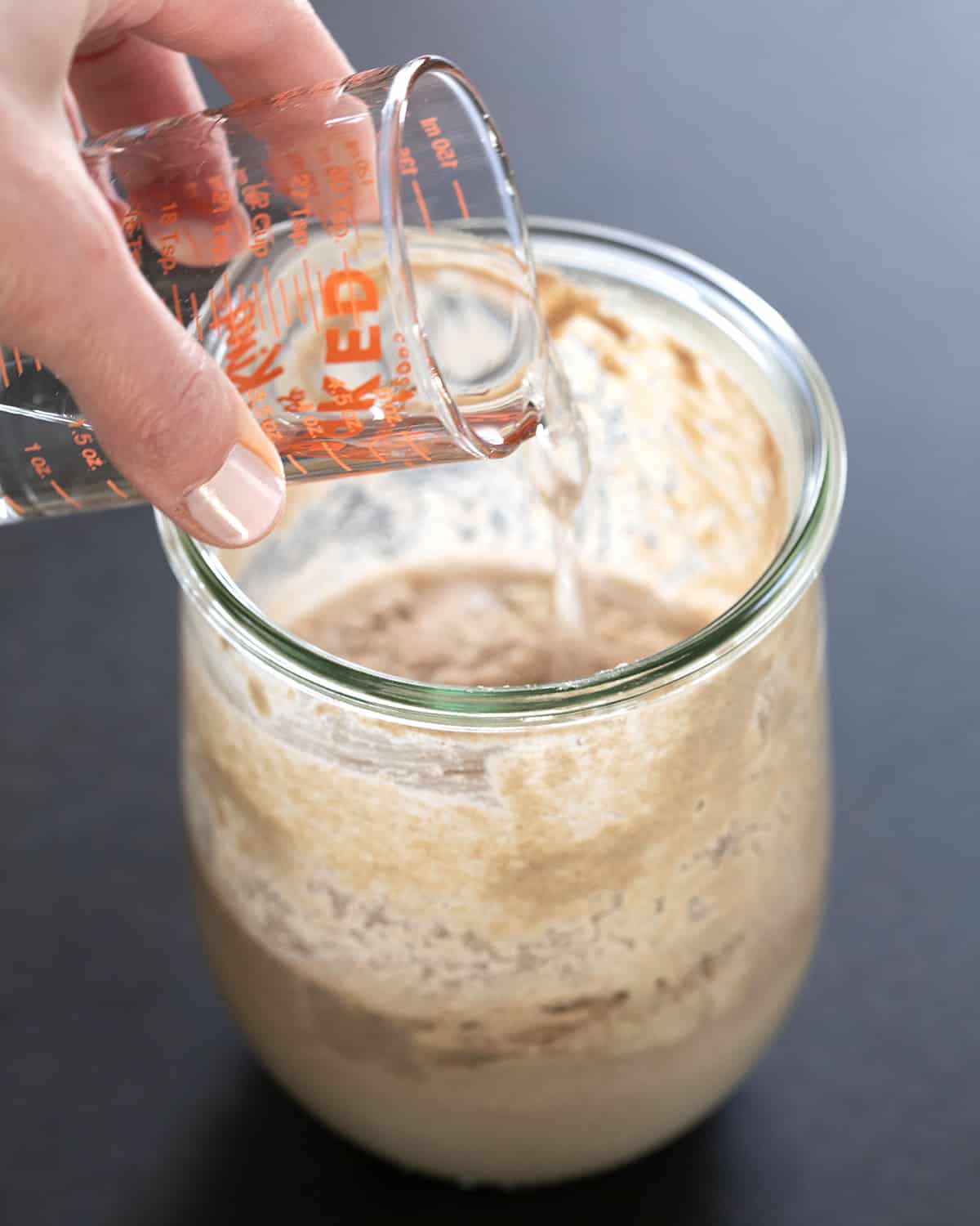hand pouring small glass jar of distilled water into sourdough starter