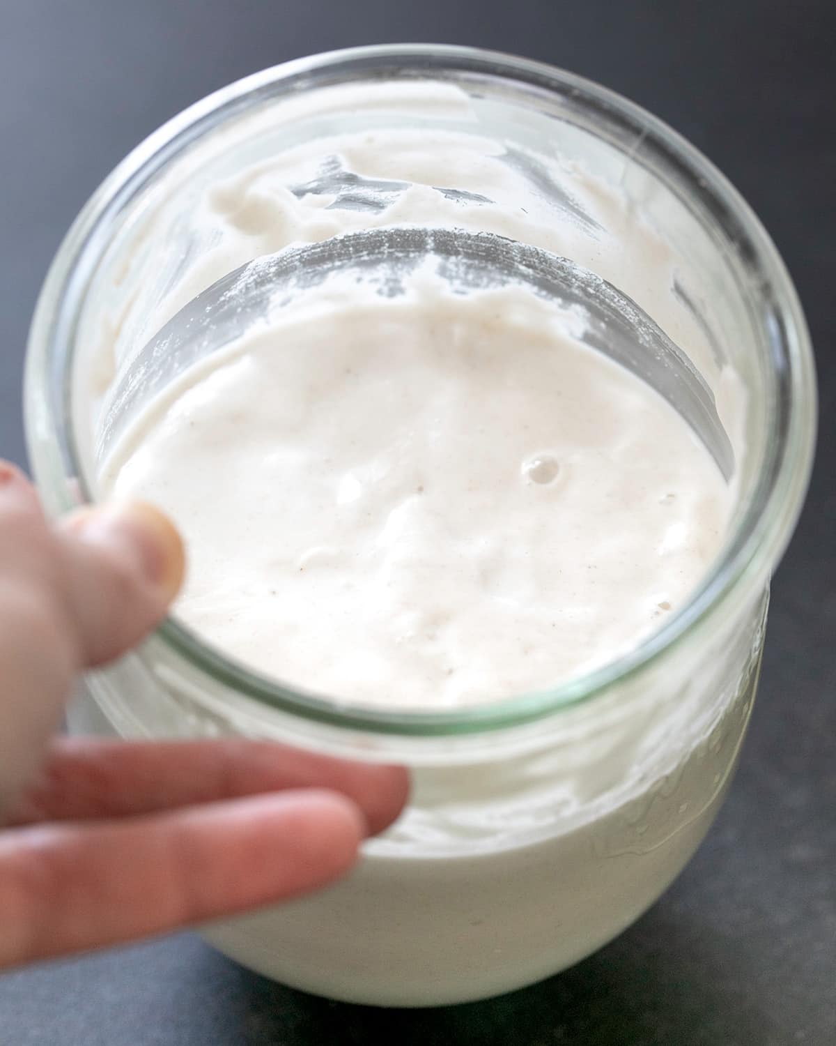 Hand holding glass jar with foamy very light brown liquid showing a rise line after the starter rose overnight.