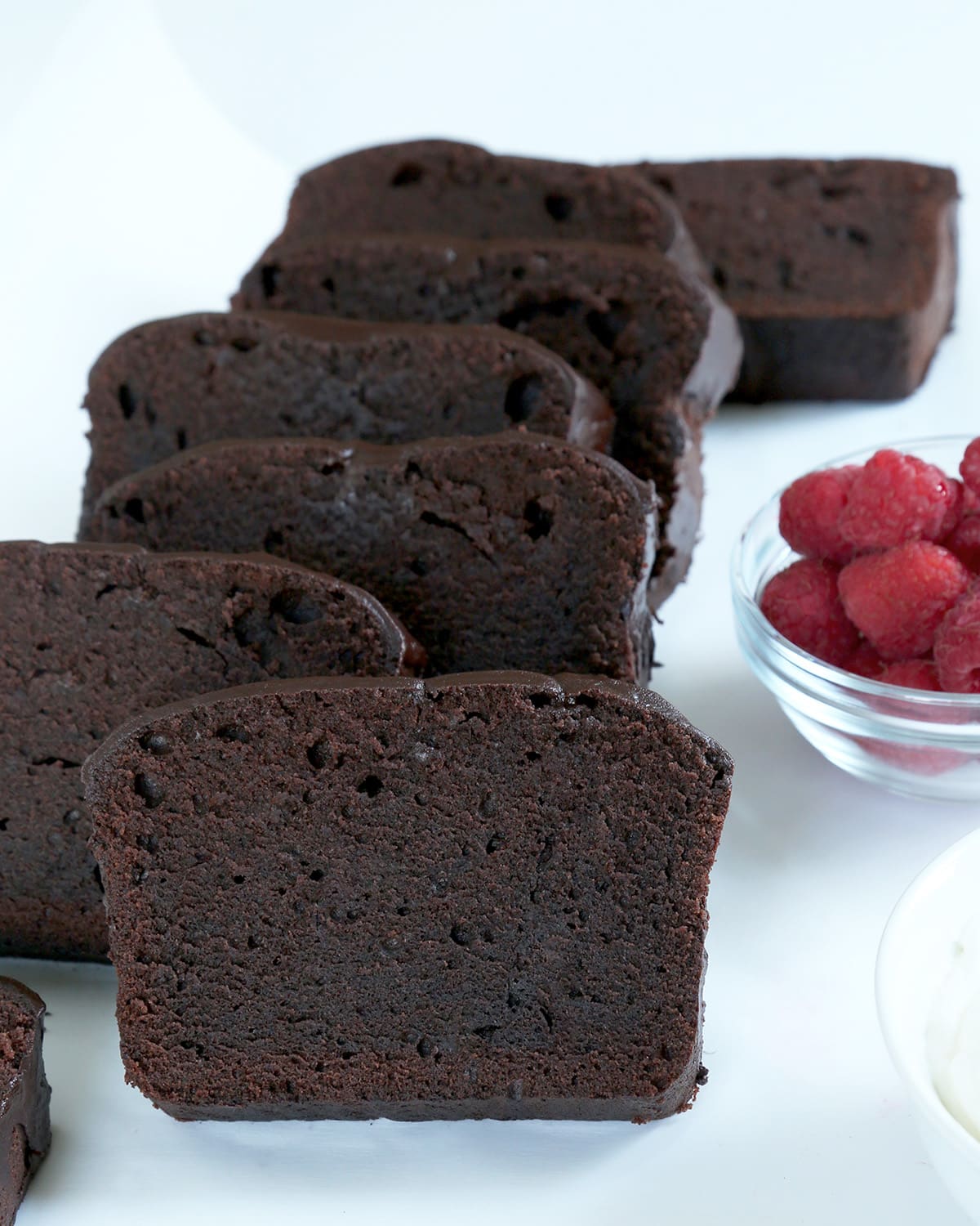 Slices of chocolate pound cake leaning against each other on a white surface with a bowl of raspberries in back