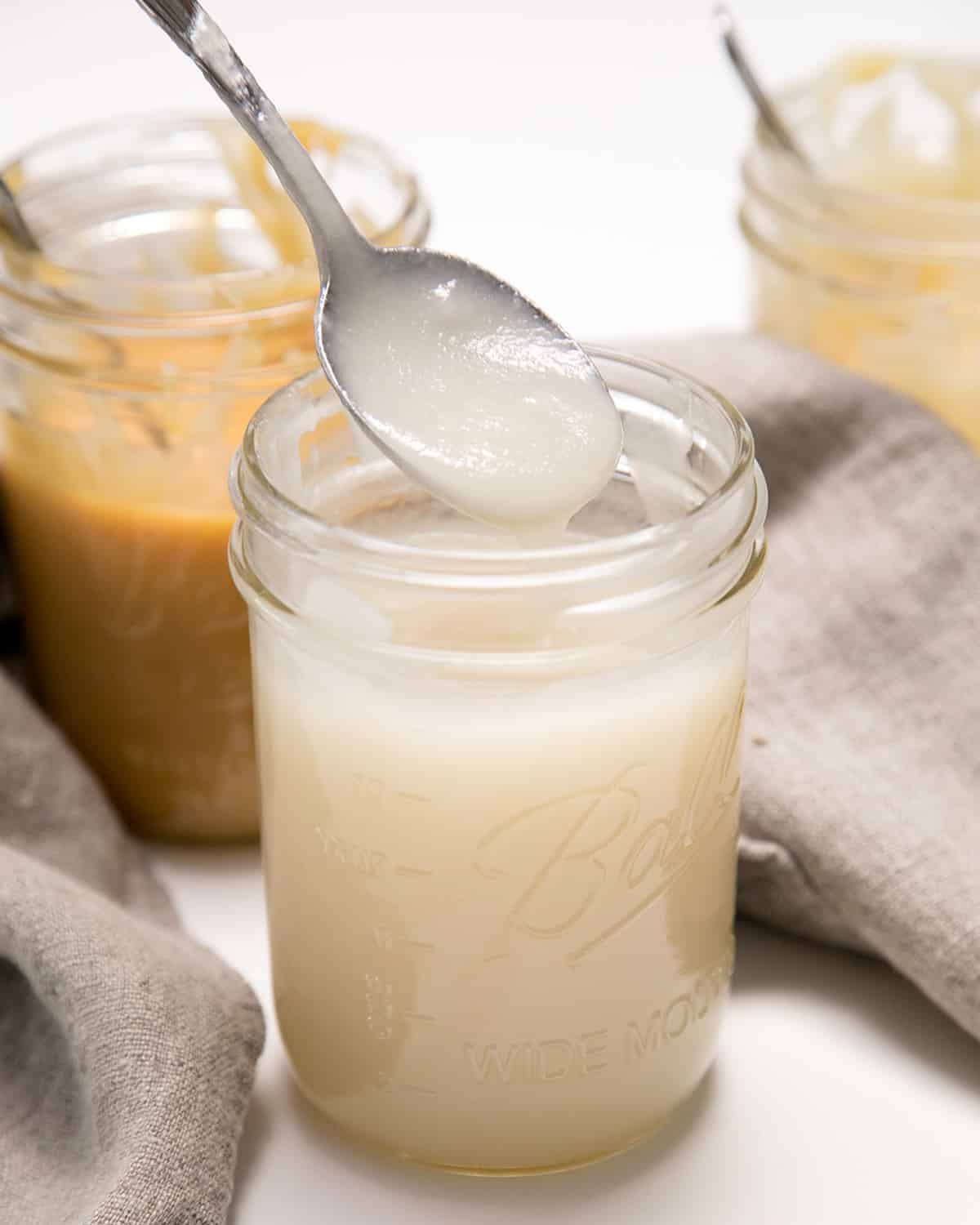 A spoon lifts a thick white mixture above a glass jar, with other jars blurred in the background.