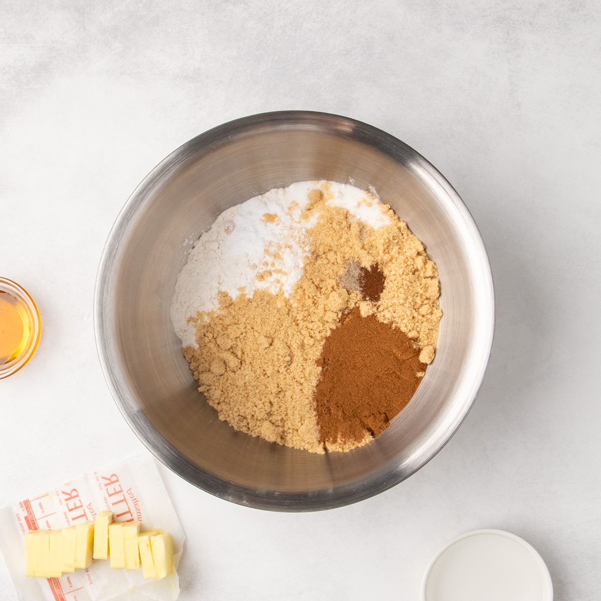 White flour, brown sugar, cinnamon, and other dry ingredients sit in separate piles in a large metal mixing bowl, with sliced butter nearby.