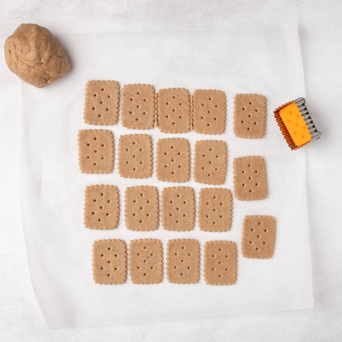 Rows of unbaked rectangular cookies with scalloped edges and small holes sit on parchment paper beside a ball of dough and a rectangular cutter.