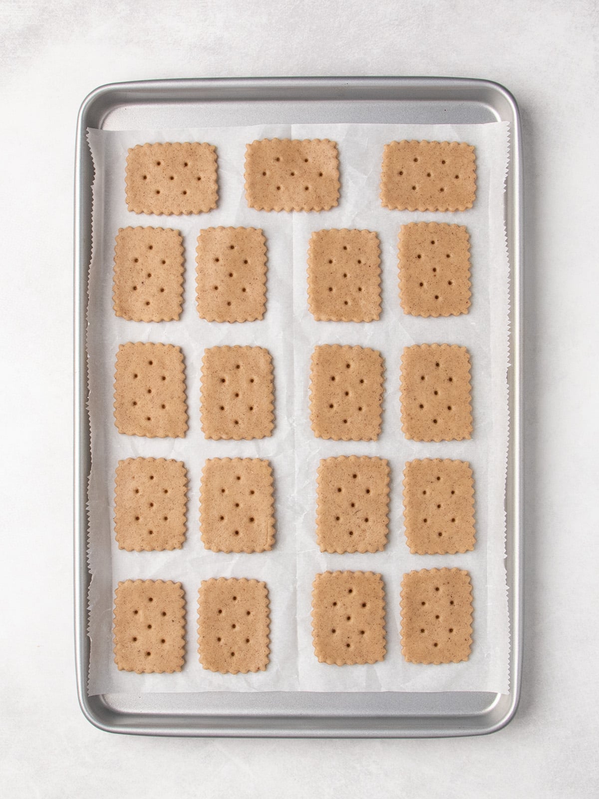 Raw rectangular cookies with scalloped edges and small holes sit in rows on a parchment-lined baking sheet.