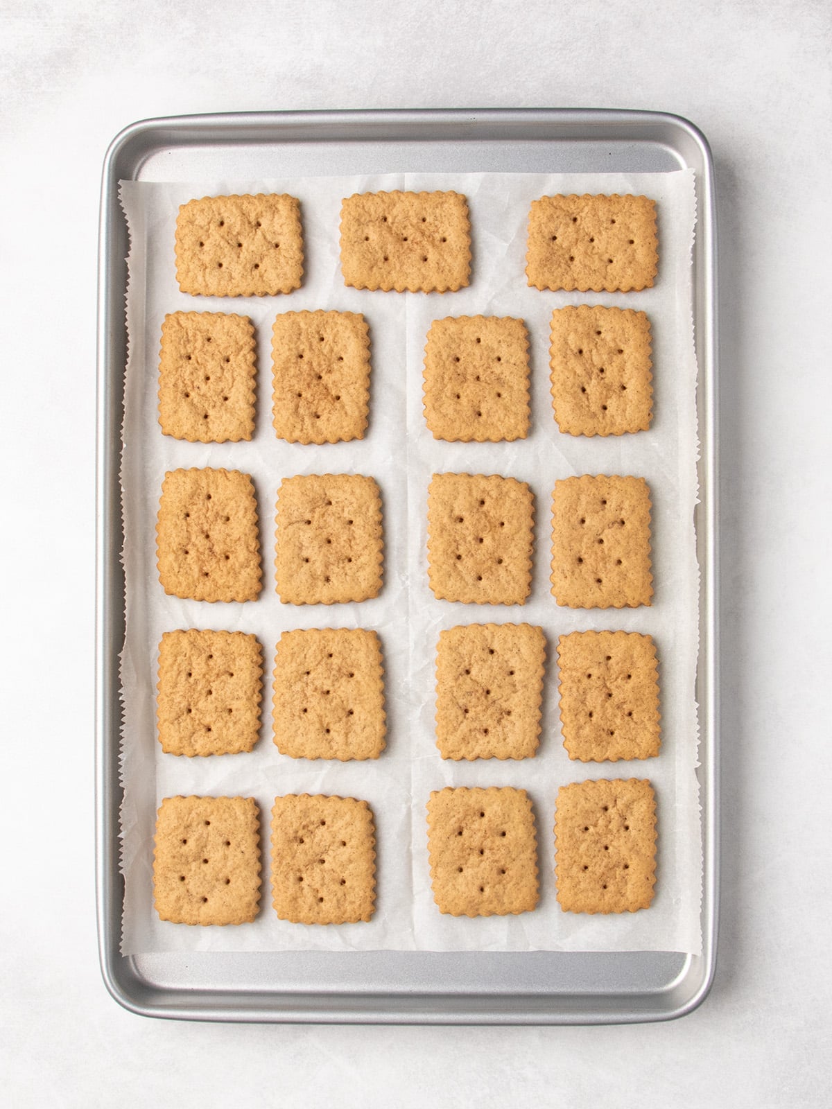 Baked light brown rectangular cookies with scalloped edges and small holes sit in rows on a parchment-lined baking sheet.