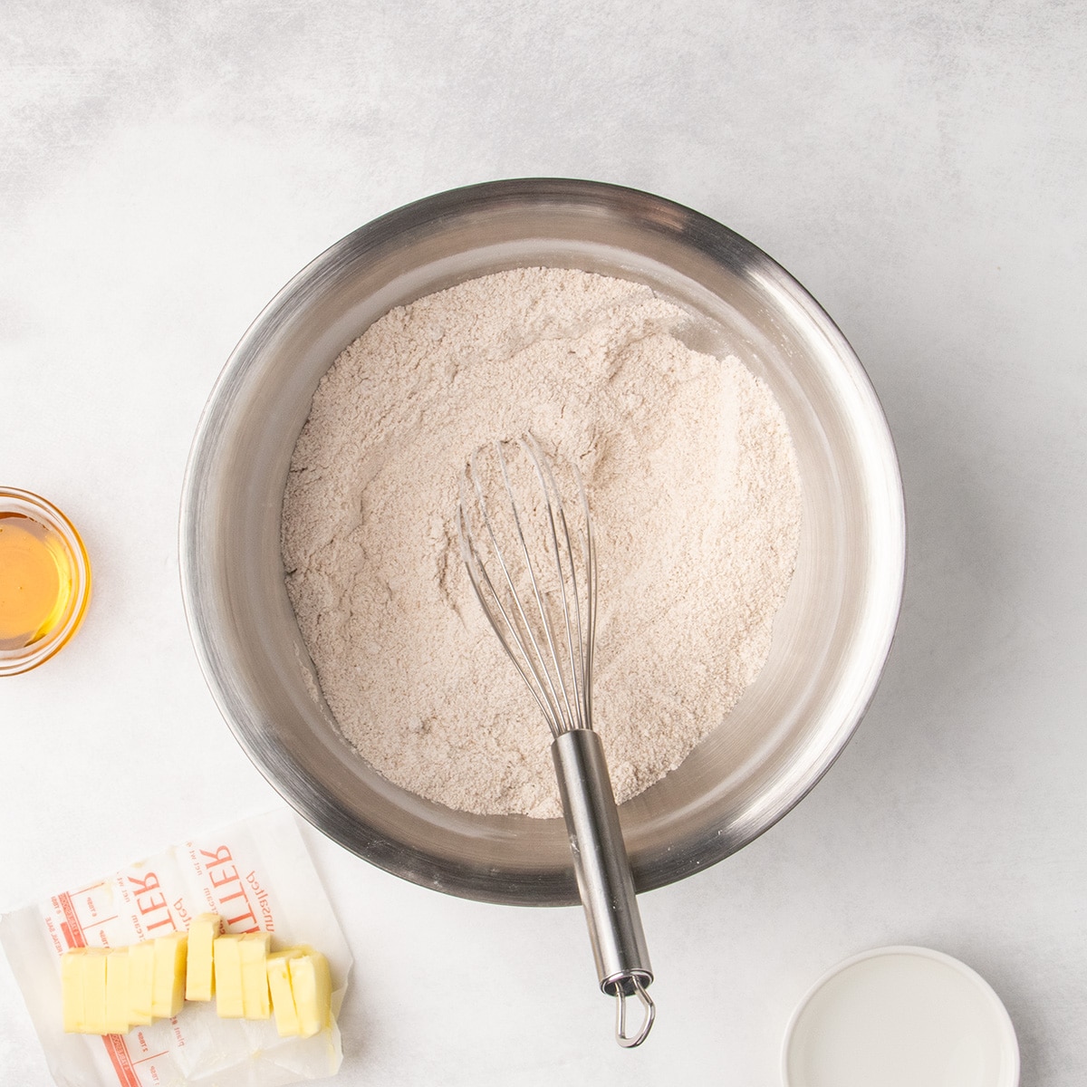 A whisk rests in the combined light brown dry ingredients in a large metal mixing bowl, with sliced butter and small bowls nearby.