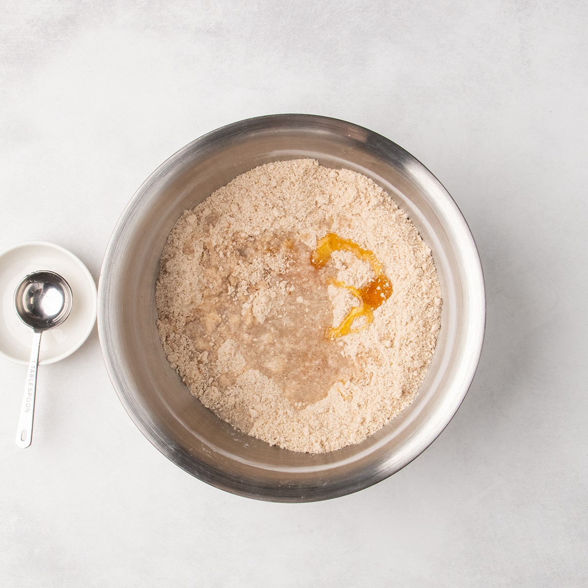 Liquid ingredients sit in the center of a crumbly light brown mixture in a large metal mixing bowl.