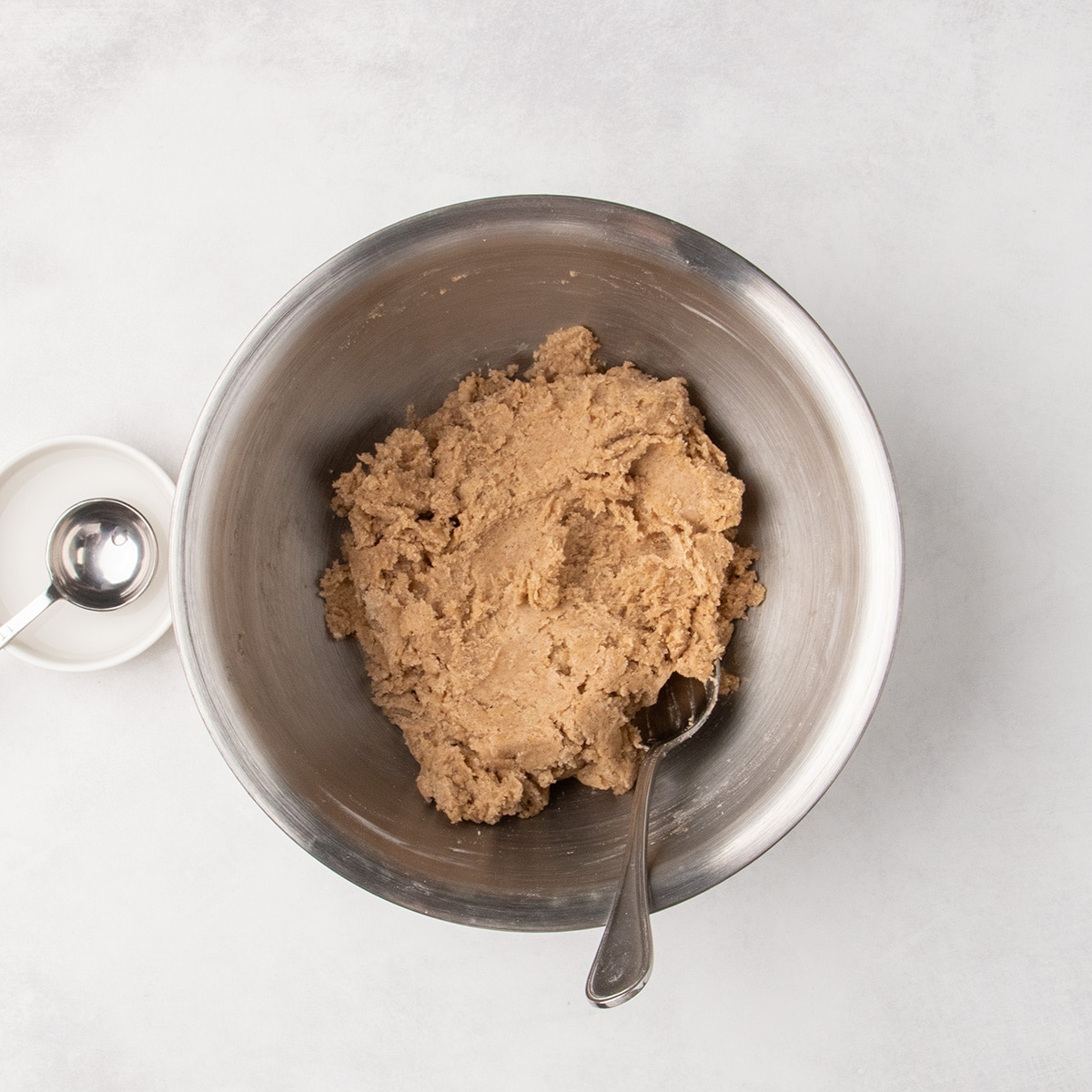 A spoon rests in thick light brown raw cookie dough in a large metal mixing bowl.