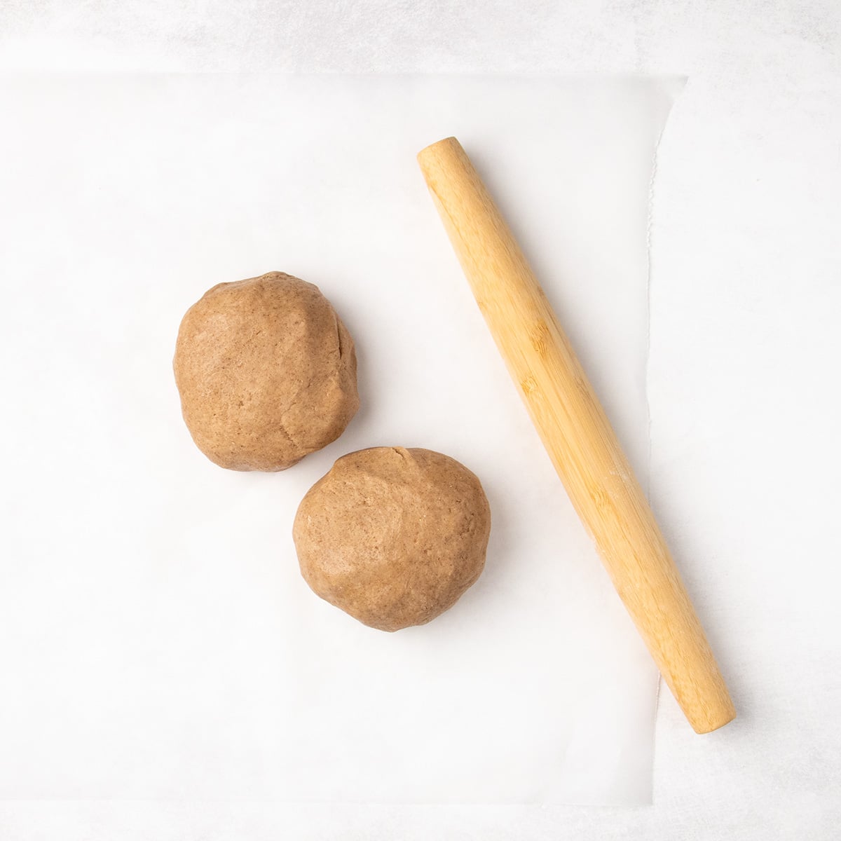 Two balls of light brown raw cookie dough sit on parchment paper beside a wooden rolling pin.