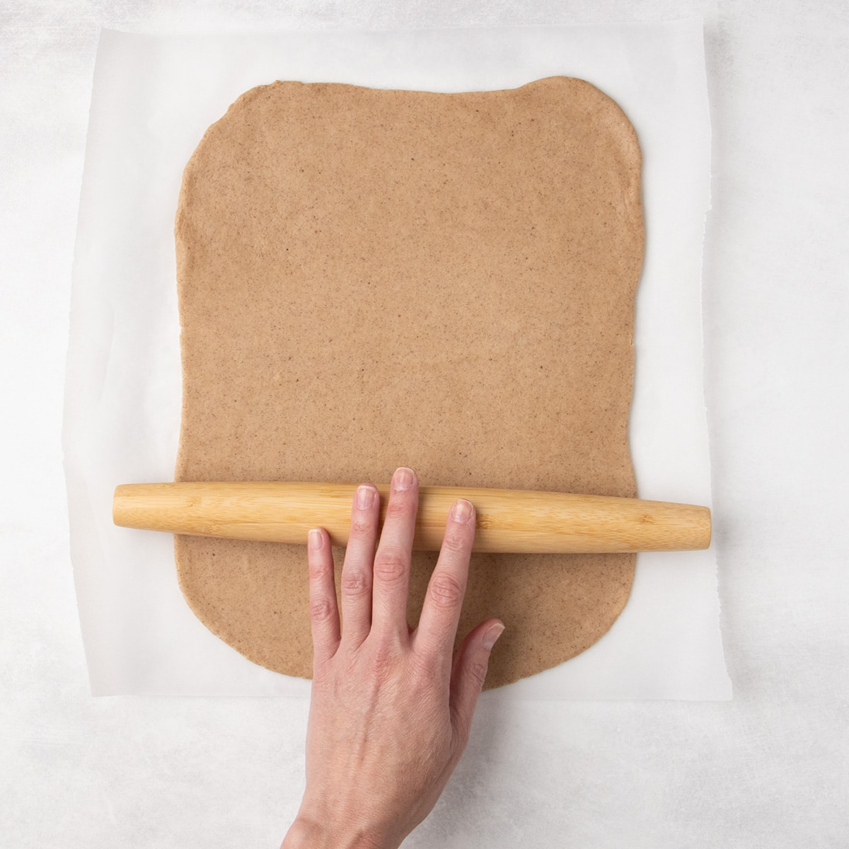 A hand rolls a large rectangular sheet of light brown raw cookie dough on parchment paper with a wooden rolling pin.