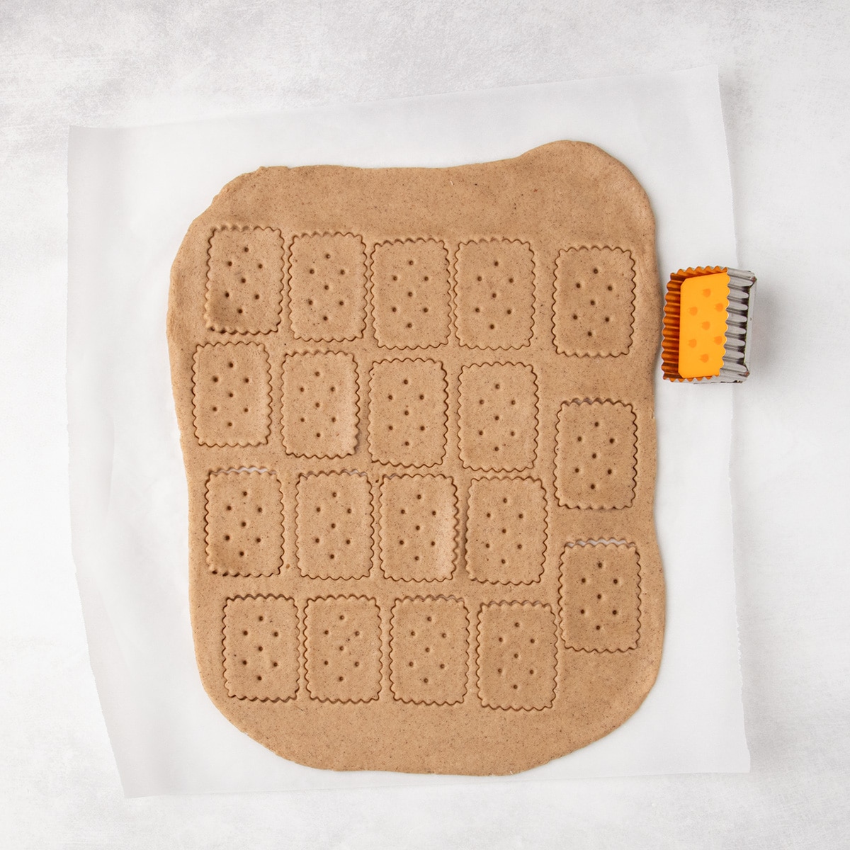 15 rectangular raw cookies with scalloped edges and small holes are cut into a sheet of rolled brown dough, with a rectangular cutter beside it.