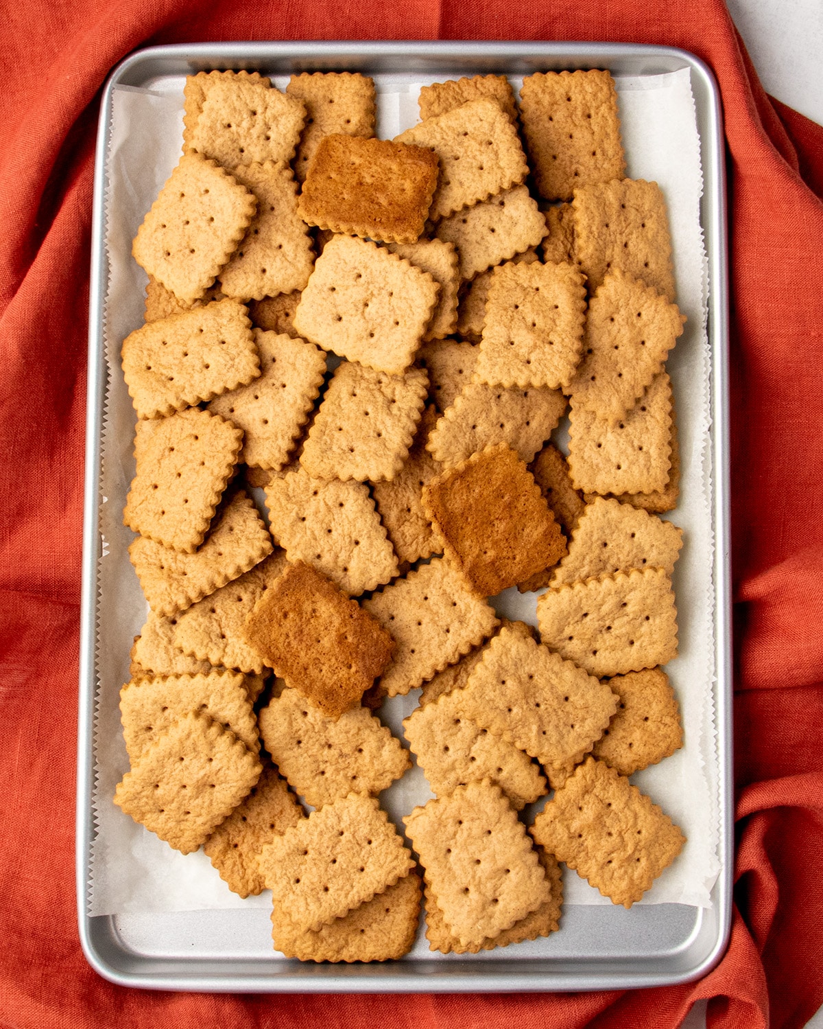 Overhead image of a parchment-lined baking sheet filled with light brown baked rectangular scalloped cookies, resting on a red cloth.