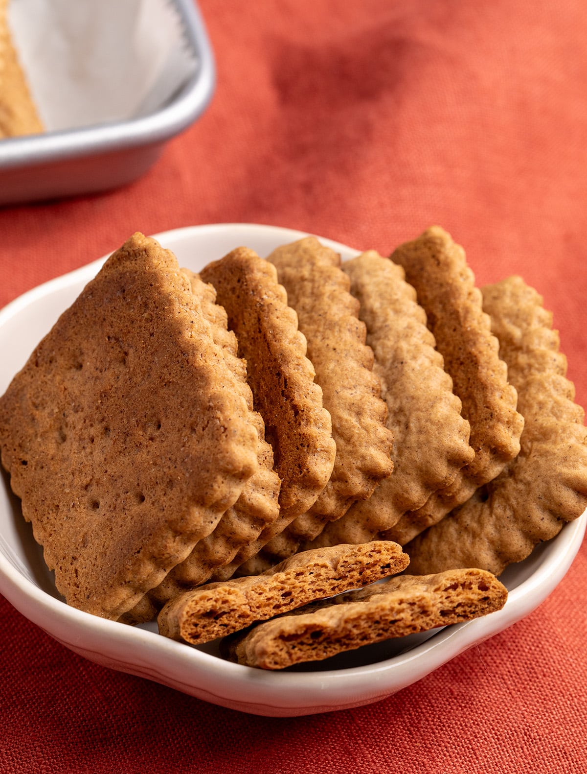 Rectangular scalloped cookies stand upright on a small white flat bowl, with two broken cookies showing their crispy interior.