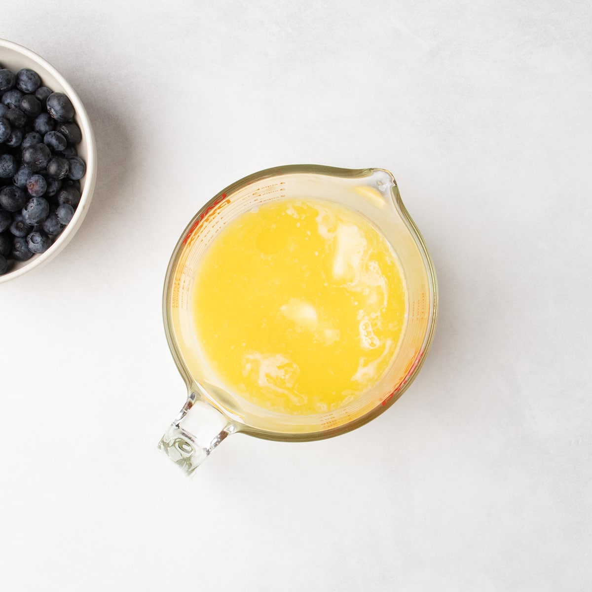 Melted yellow liquid sits in a glass measuring cup beside a bowl of blueberries.