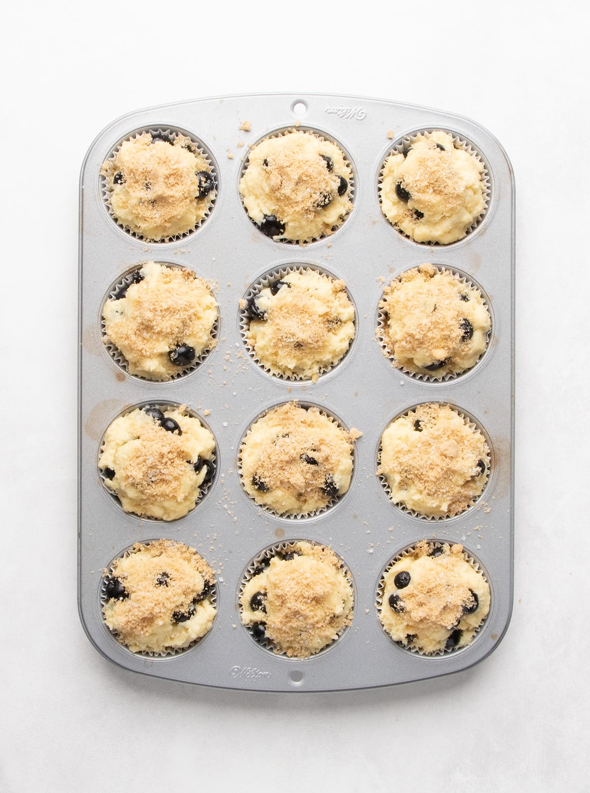 Twelve unbaked blueberry muffins topped with light brown sugar sit in a metal muffin pan.