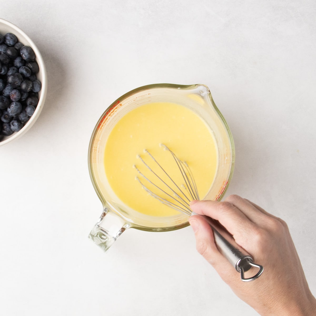 A hand whisks a pale yellow liquid in a glass measuring cup beside a bowl of blueberries.