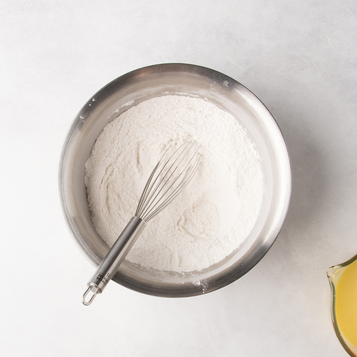 A whisk rests in white powdered ingredients in a stainless steel mixing bowl.