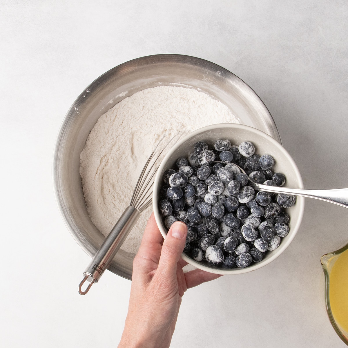 A hand holds a bowl of blueberries coated in white powder over a bowl of dry ingredients.