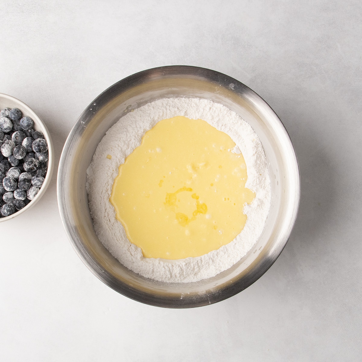 Pale yellow liquid sits in the center of white dry ingredients in a stainless steel mixing bowl.