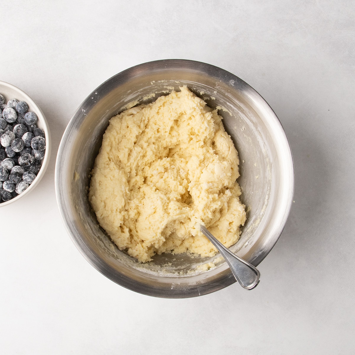 A spoon rests in thick pale yellow batter in a stainless steel mixing bowl beside a bowl of coated blueberries.