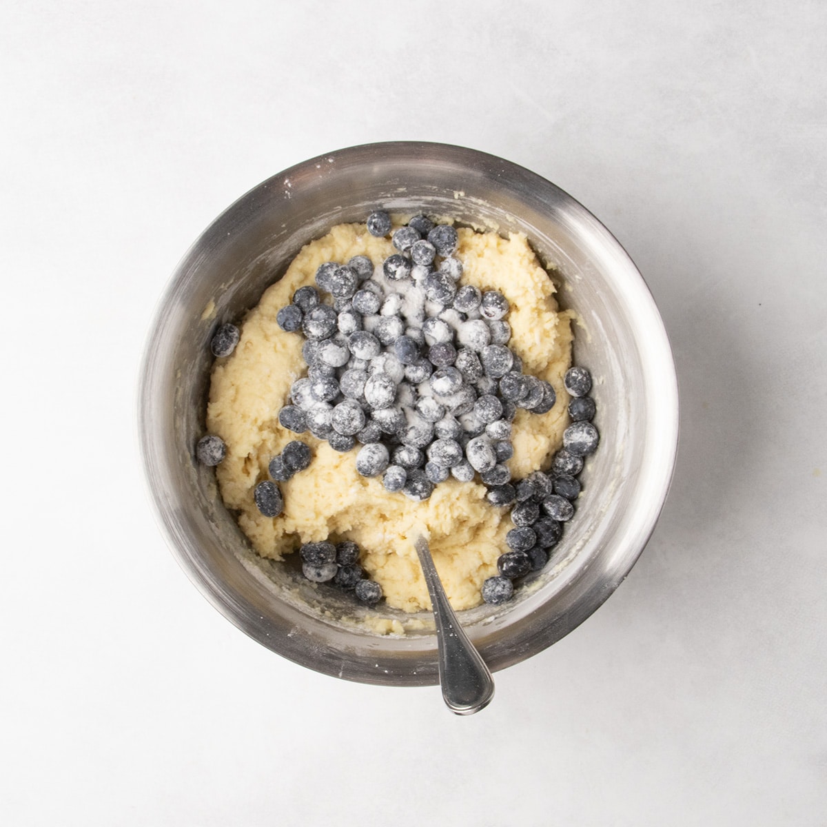 Blueberries coated in white powder sit on top of thick pale yellow batter in a stainless steel mixing bowl.