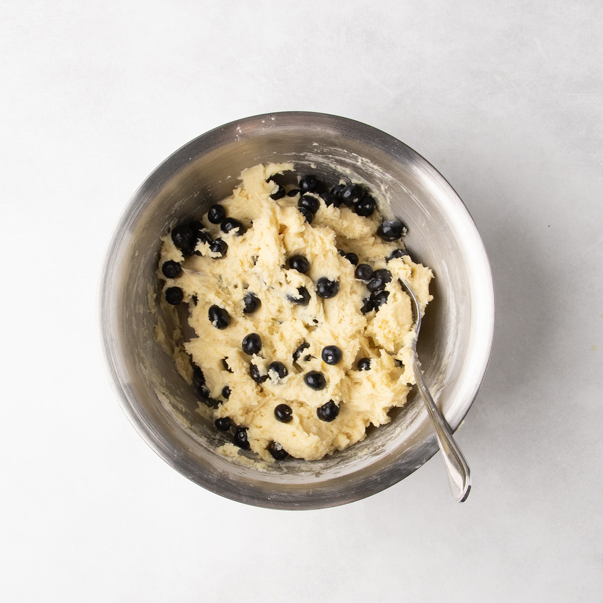 Blueberries are mixed throughout thick pale batter in a stainless steel mixing bowl with a spoon resting inside.