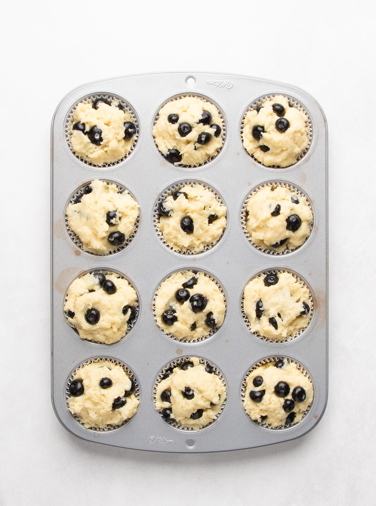 Twelve paper liners filled with pale yellow raw batter and blueberries sit in a metal muffin pan.