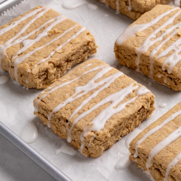 closeup image of 5 square oat-speckled gluten free breakfast bars with white glaze drizzle, resting on a parchment-lined baking sheet.