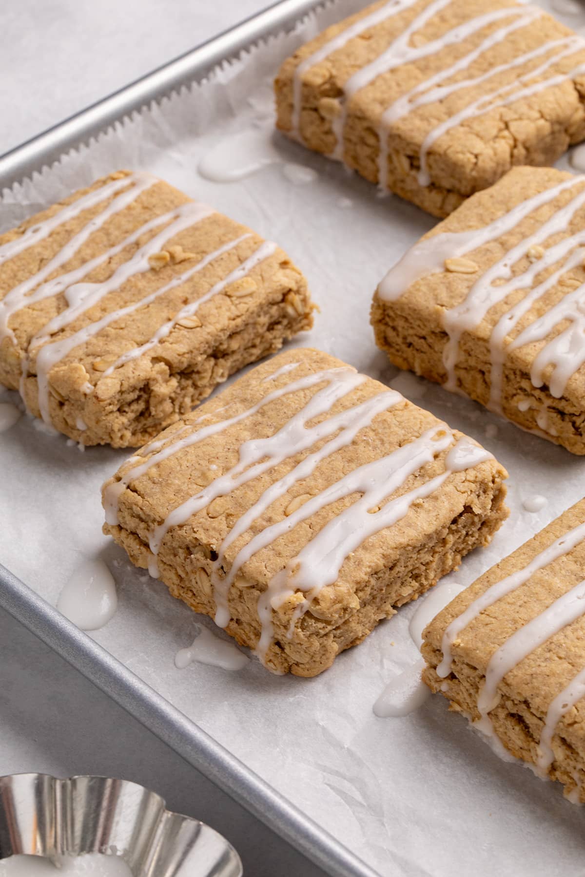 Square gluten free breakfast bars with white glaze rest on a parchment-lined baking sheet beside a small dish of glaze.