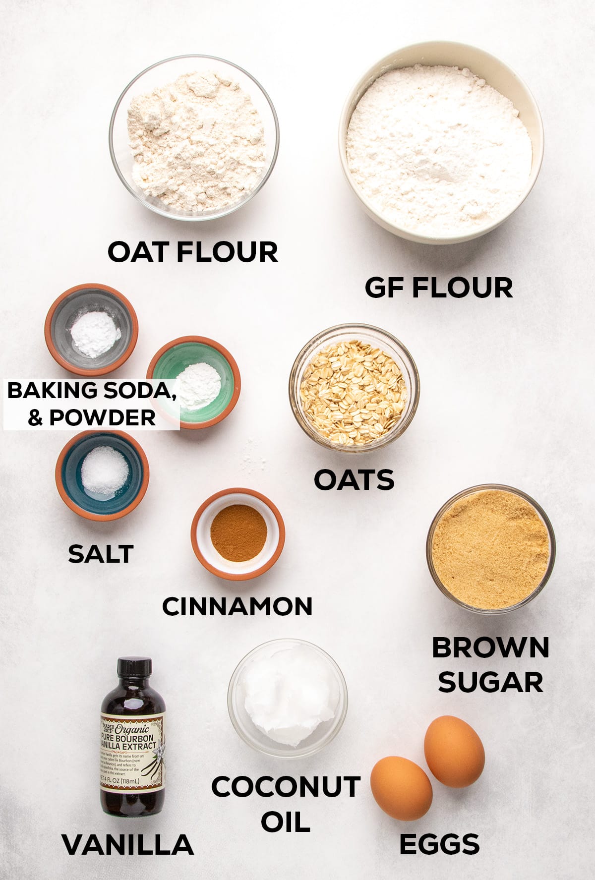 Oat flour, gluten free flour, baking soda, baking powder, salt, oats, cinnamon, brown sugar, vanilla, coconut oil, and two eggs are arranged on a light surface with text labels.