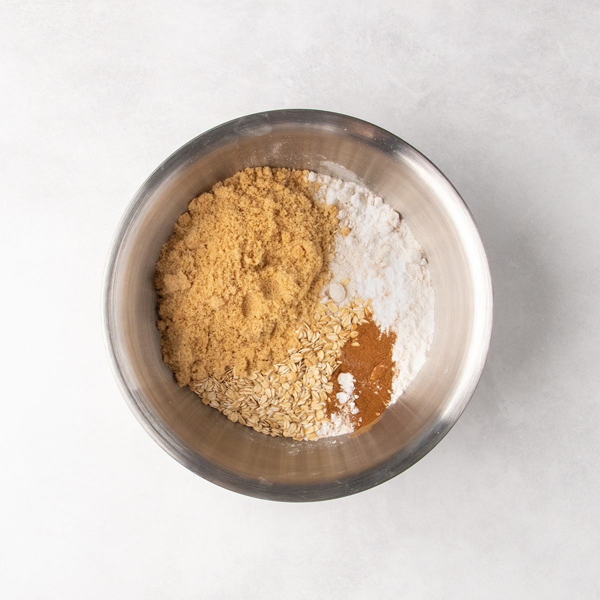 Light brown sugar, white gluten free flour, rolled oats, cinnamon, and white powders sit in separate piles in a stainless steel mixing bowl.