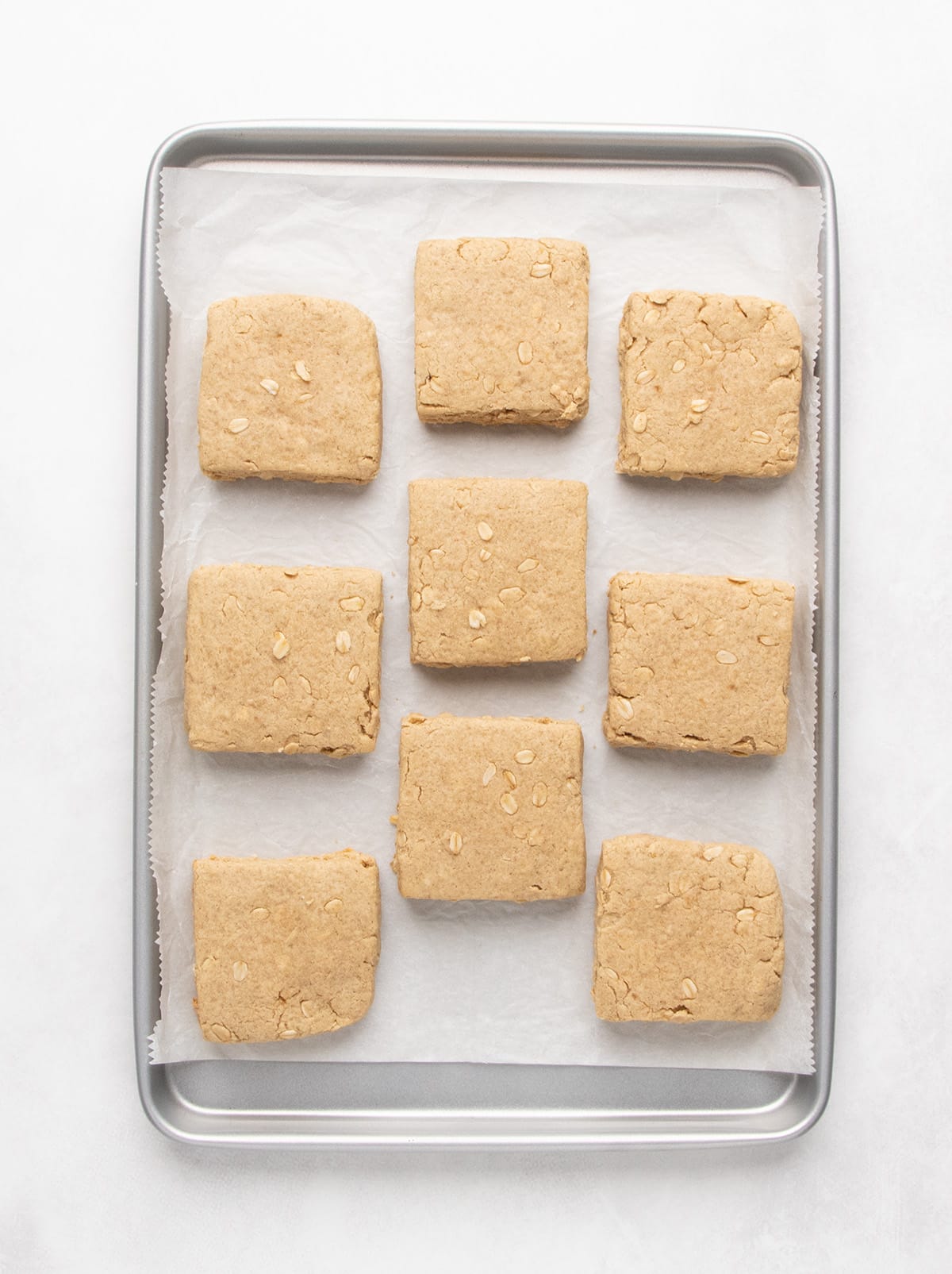 Nine baked square bars with lightly browned surfaces rest on a parchment-lined metal baking sheet.