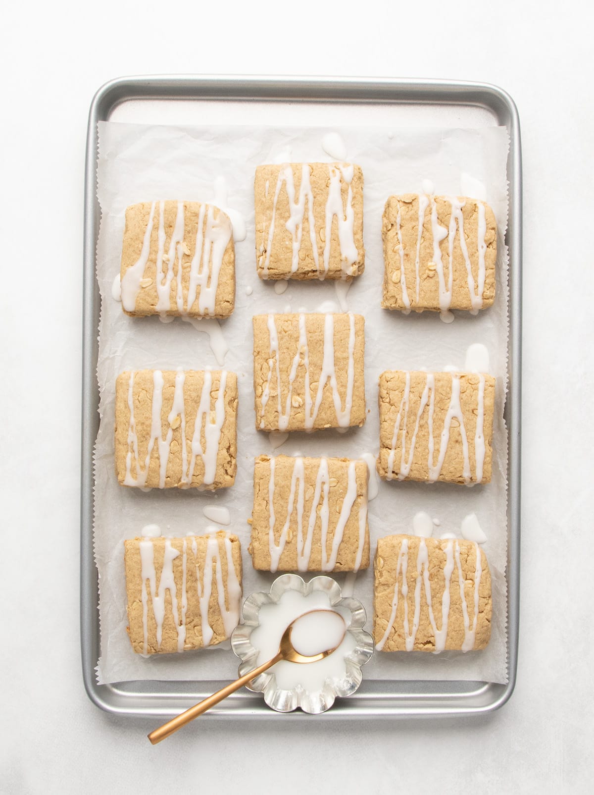 Nine baked square breakfast bars with white glaze drizzled over the tops rest on a parchment-lined baking sheet beside a small dish and spoon.