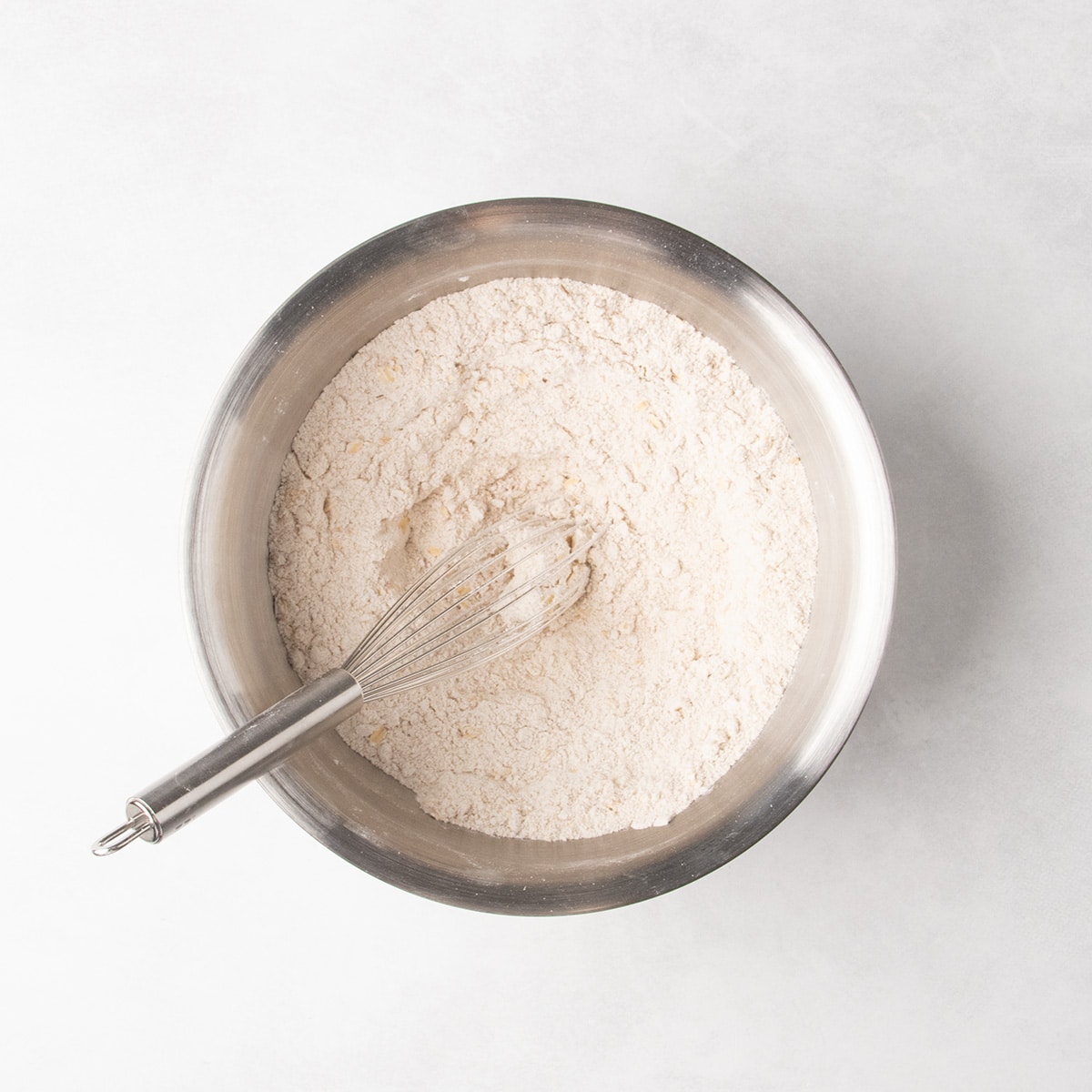 A whisk rests in pale beige dry ingredients combined in a stainless steel mixing bowl.