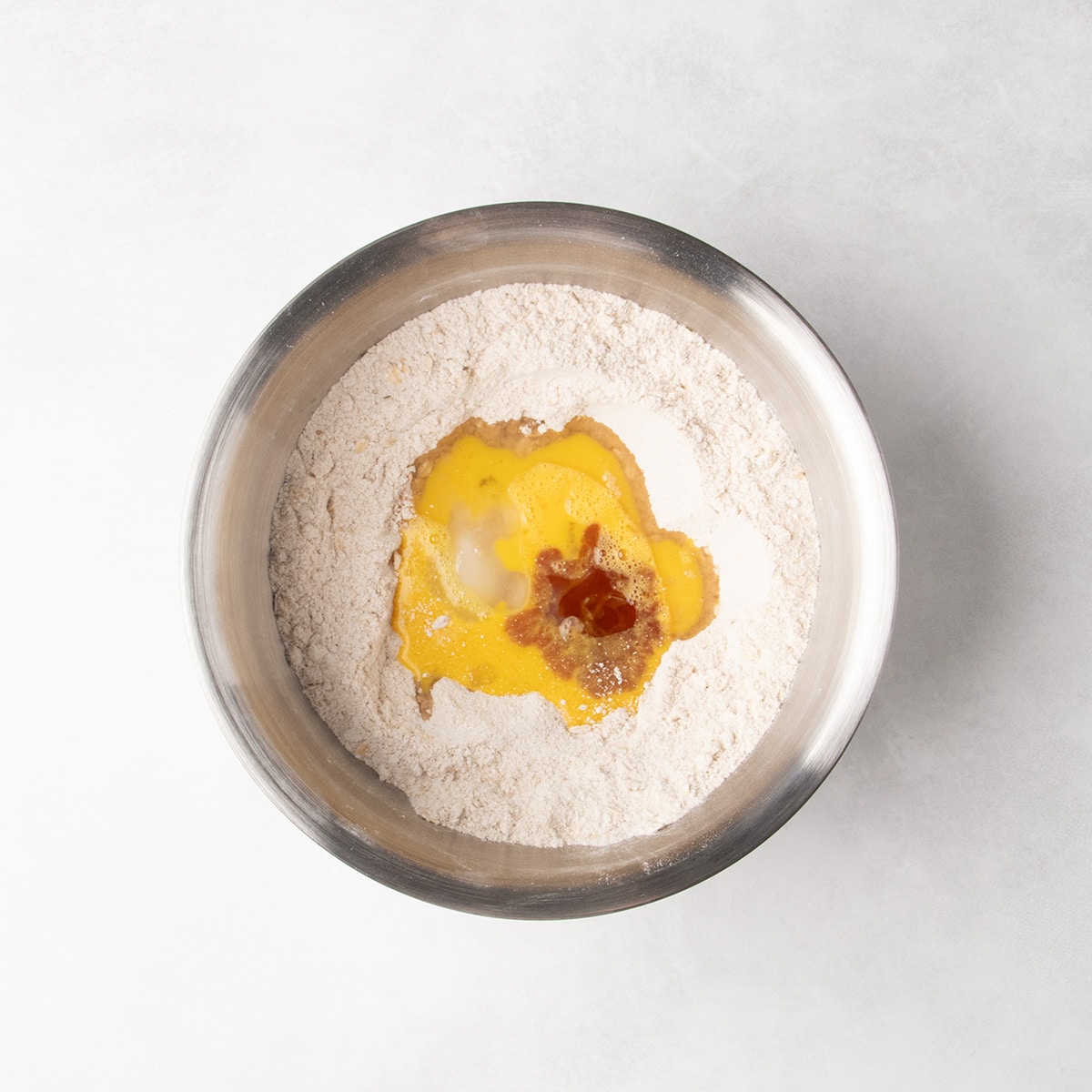 Yellow liquid, white liquid, and brown liquid sit in the center of the dry ingredients in a stainless steel mixing bowl.