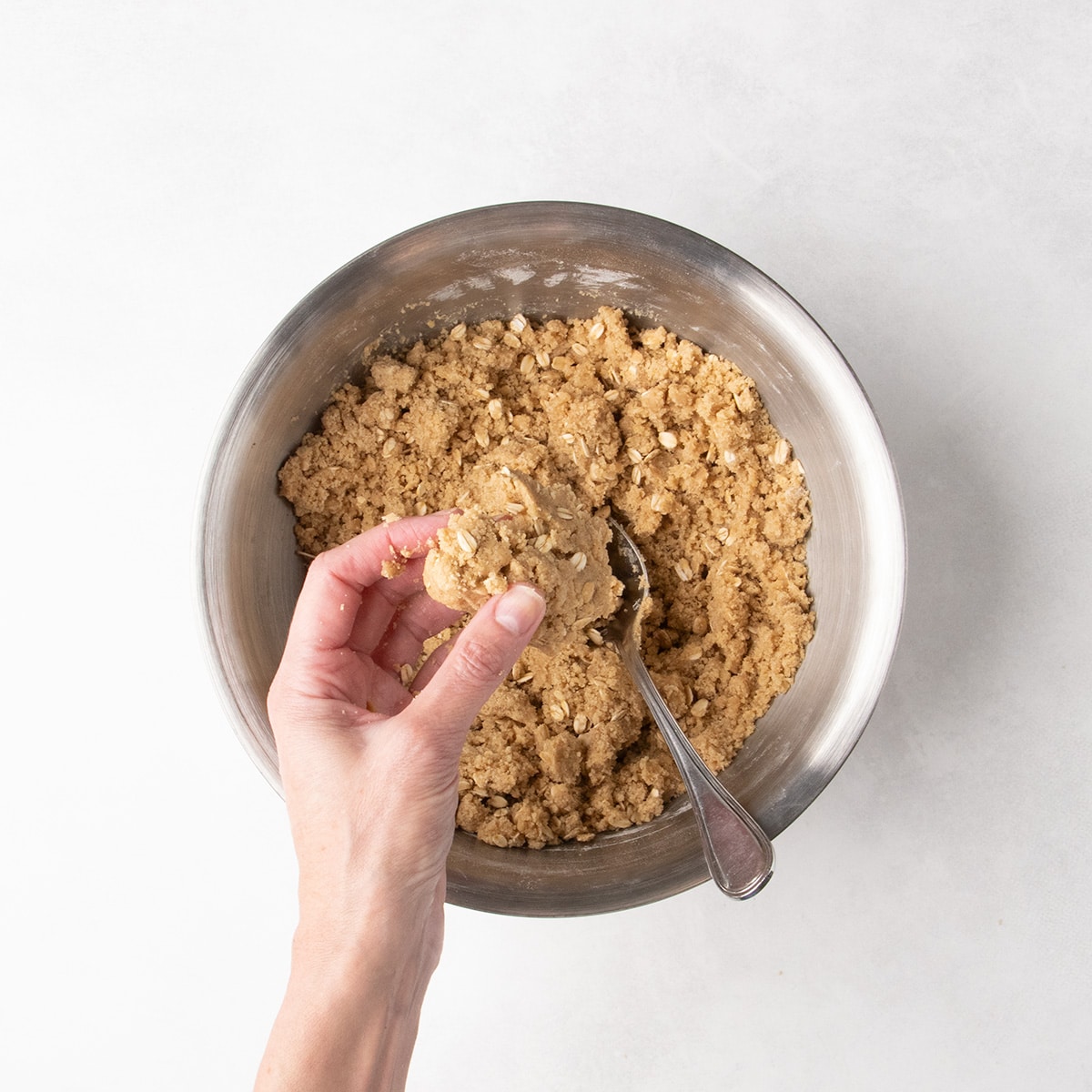 A hand holds a clump of oat dough over a stainless steel mixing bowl with a spoon resting inside.