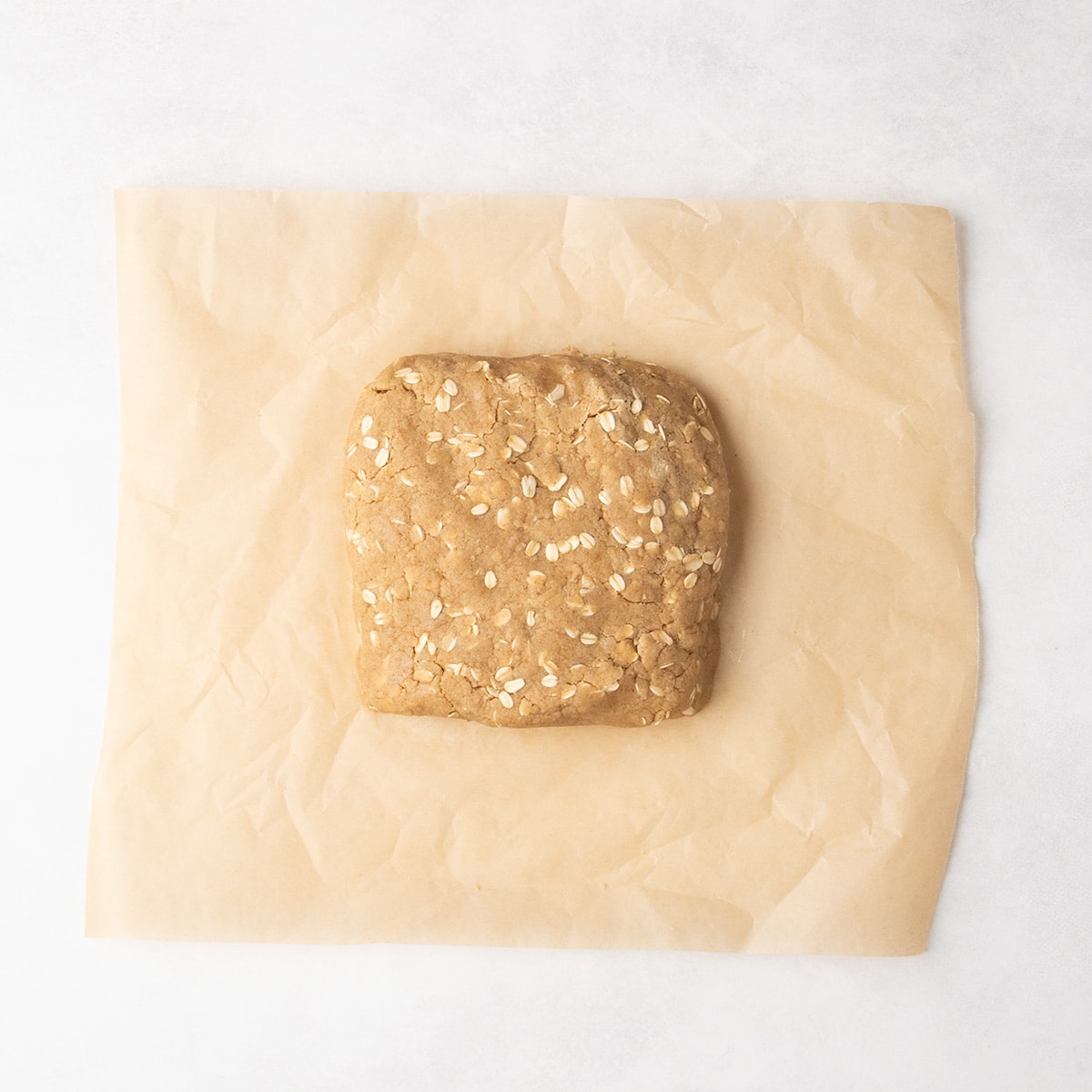 A rough square block of brown, oat-speckled dough rests on parchment paper.