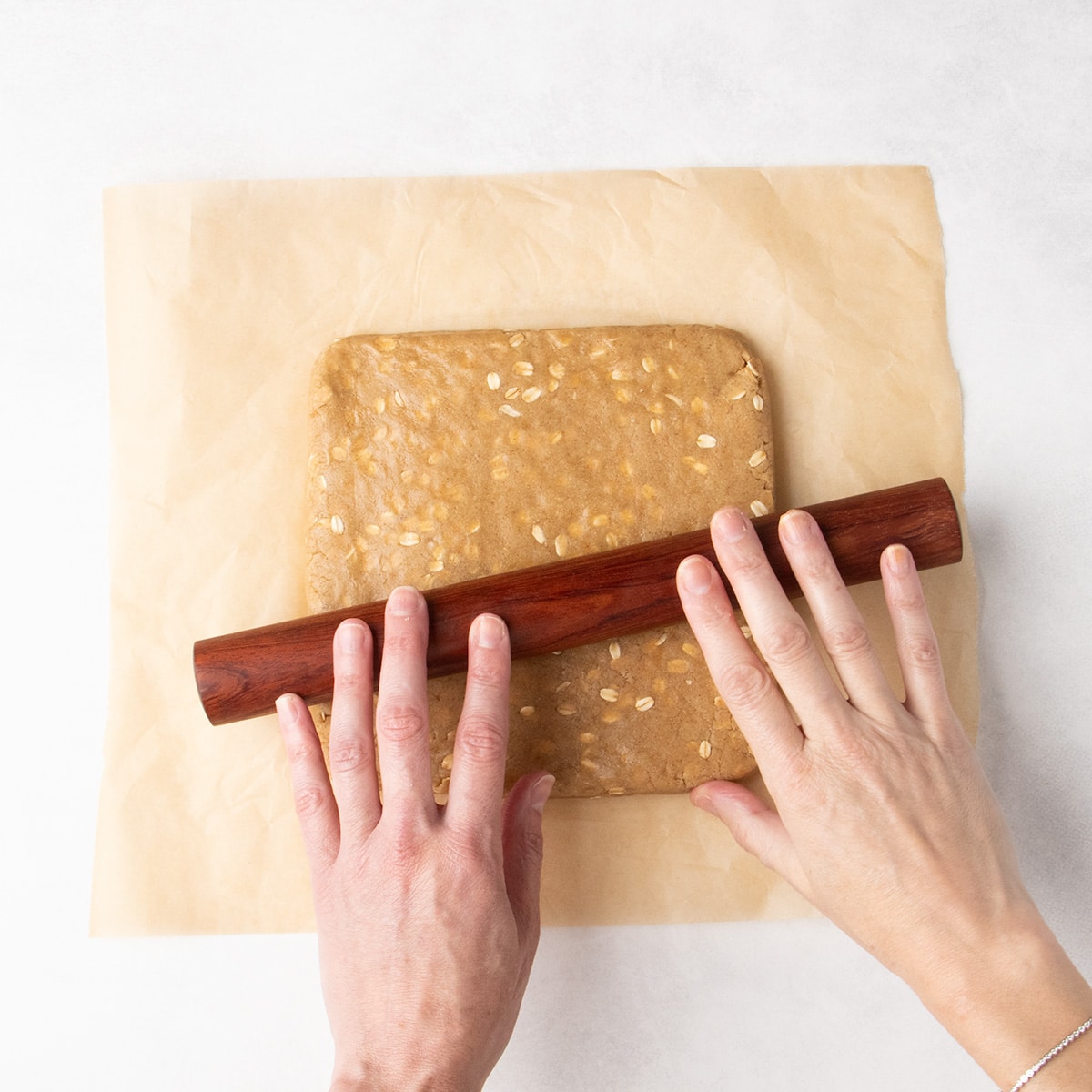 Two hands use a wooden rolling pin to flatten brown raw oat-speckled dough on parchment paper.