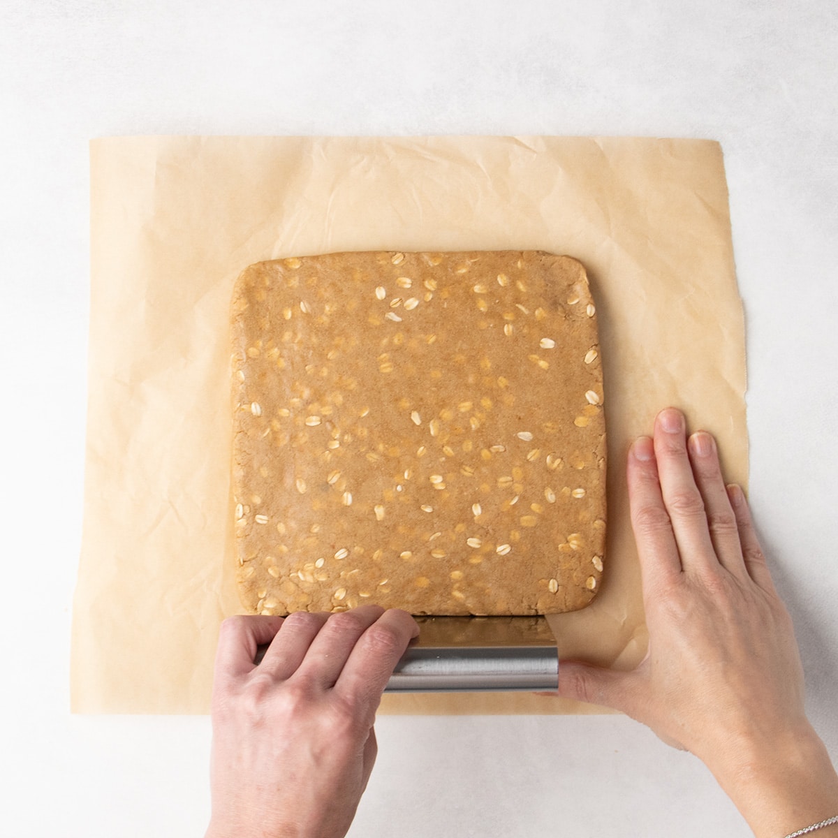 A hand holds a metal bench scraper against the lower edge of a flattened square of raw brown oat-speckled dough, while the other hand steadies the parchment paper beneath it.